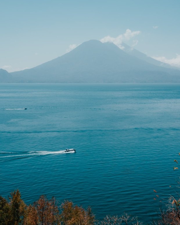 A volcano across the lake in the haze, with two small launches crossing the blue water below it.