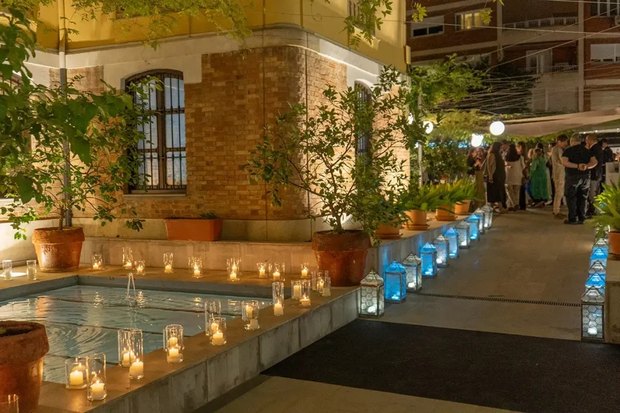 The courtyard at night: a long water basin lined with candles, lanterns along the path, and people talking under the trees.