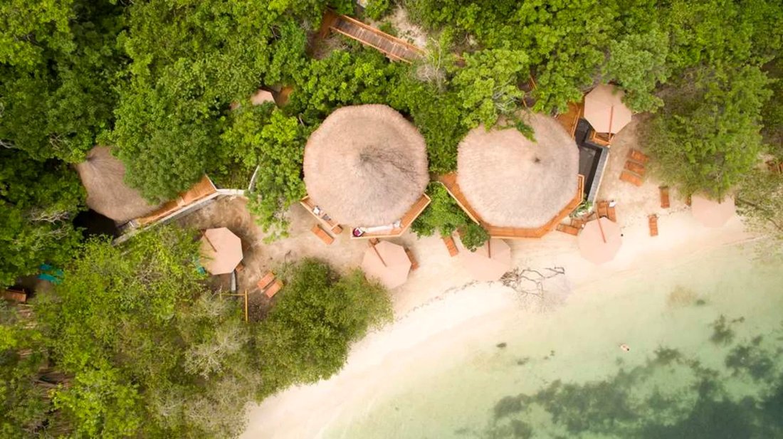 Looking straight down on the hotel: big round thatched roofs among dense green trees, a strip of pale sand and clear shallow water.