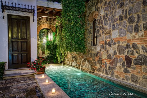 A narrow lit pool beside a stone wall with water spouts, candles along the edge and a dark wooden door at the end.