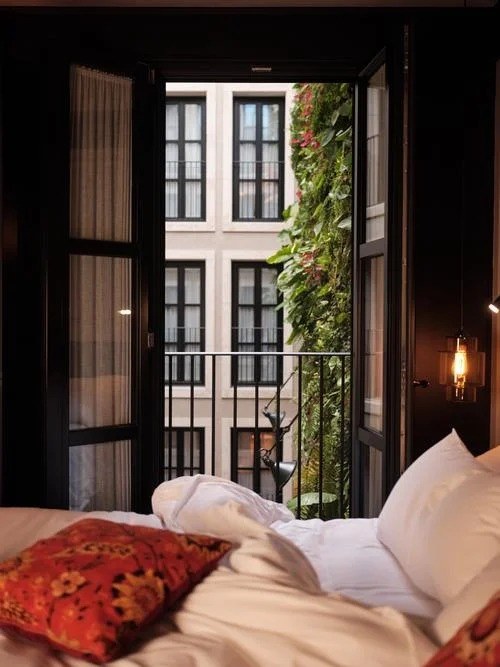 A dark-walled room, the bed in the foreground, French windows open onto a small balcony and the green wall of the courtyard.